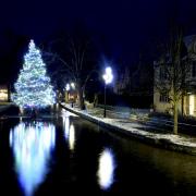 Cotswolds Christmas Scene : The world famous Bourton-On-The-Water Christmas Tree, placed and reflected in the River Windrush, is surrounded by the first sprinkling of Winter snow on the river's banks Monday 29th November 2021 in Gloucestershire. The